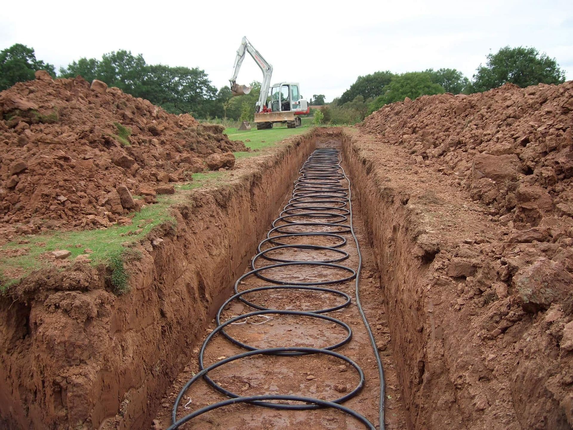 Trench with black pipes laid out for a geothermal heating system; backhoe in background.